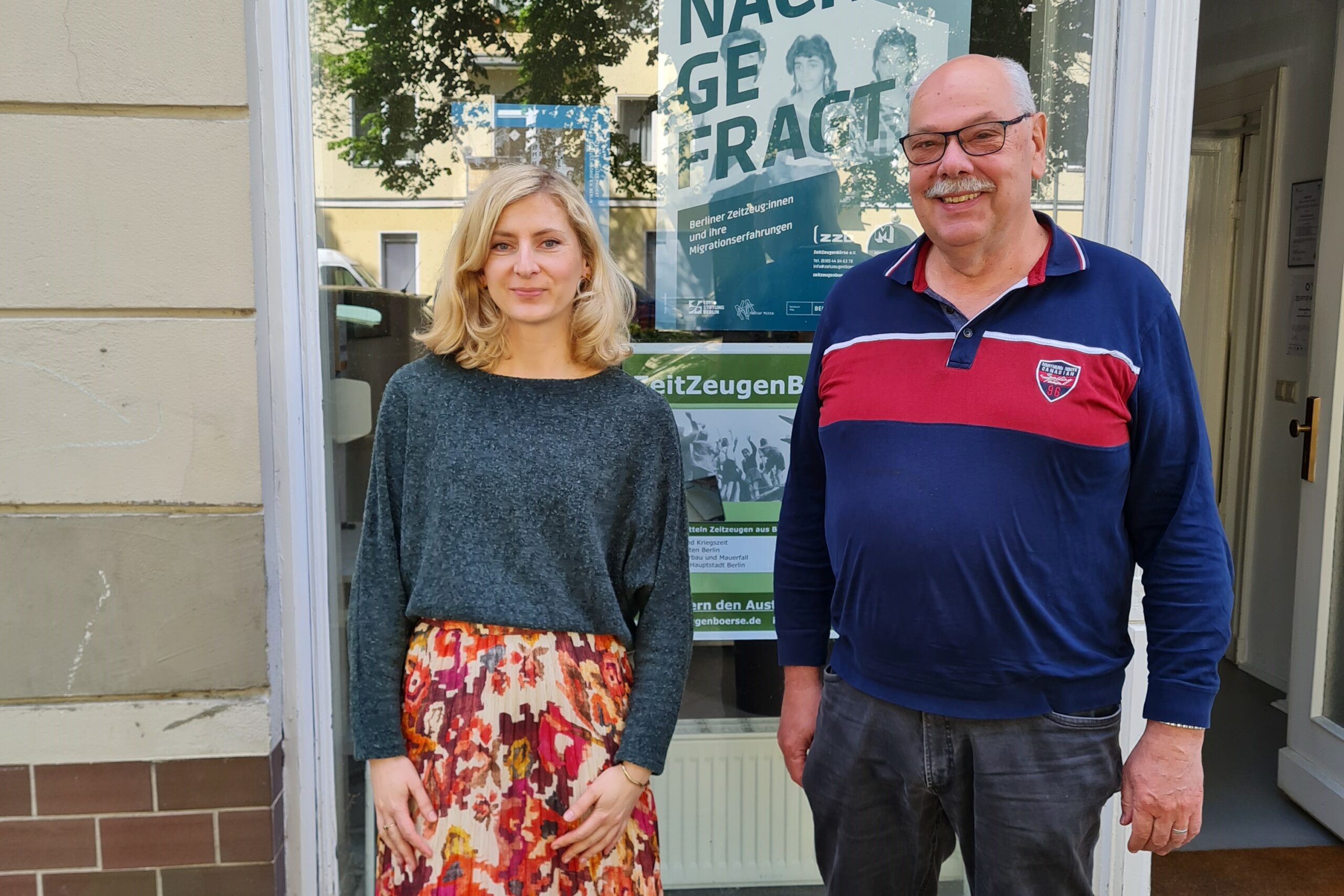 Eine junge Frau mit blonden, schulterlangen Haaren in einem grauen Pullover steht neben einem Mann in einem blau-roten Pullover. Sie stehen vor einem Ladencafé, an dem Werbeplakate für ihre Organisation hängen.