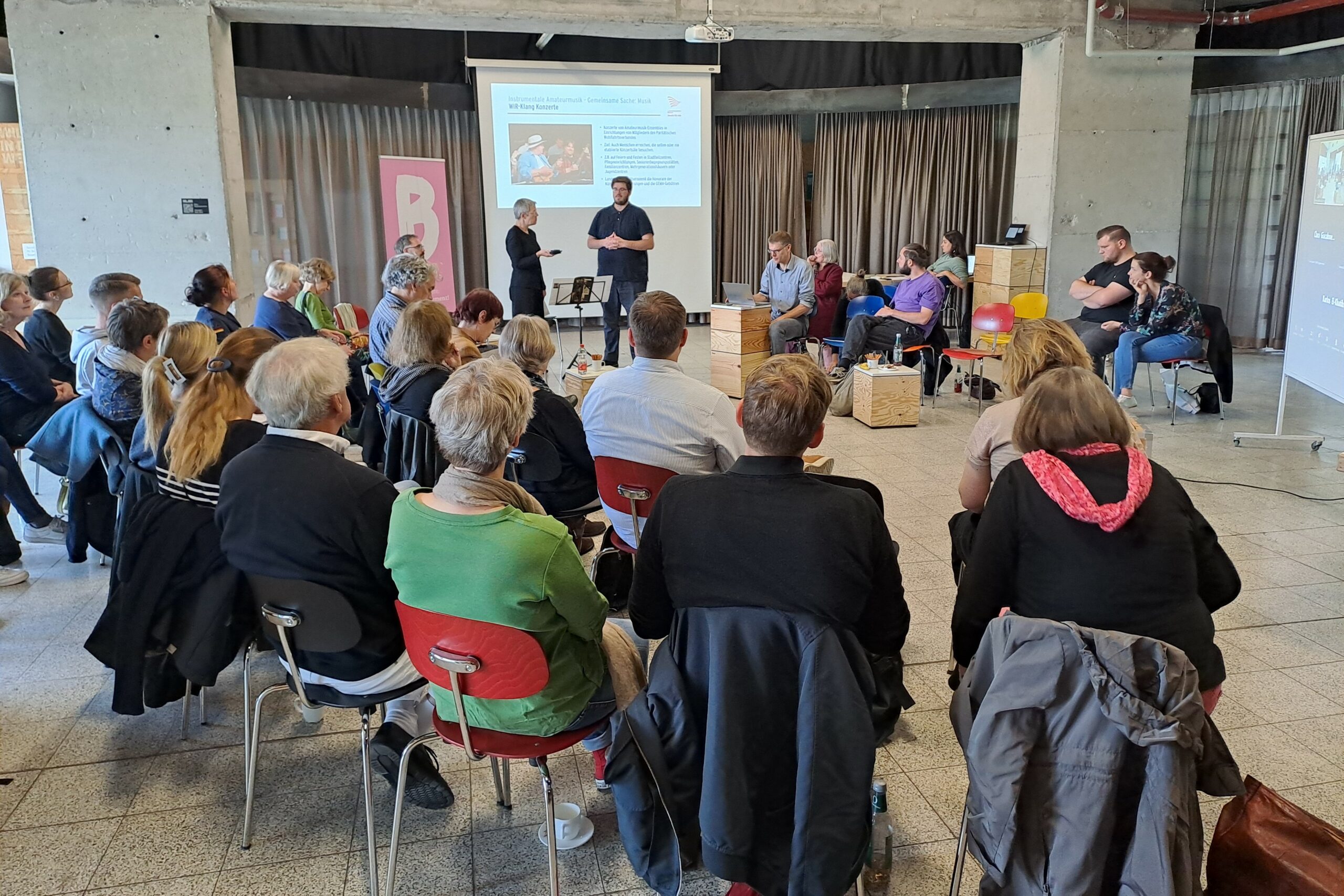 Rund 30 Teilnehmende einer Veranstaltung sitzen sich im Halbkreis gegenüber. Im Hintergrund ist eine Leinwand mit Präsentation zu sehen. Davor stellen zwei Personen ein Thema vor.