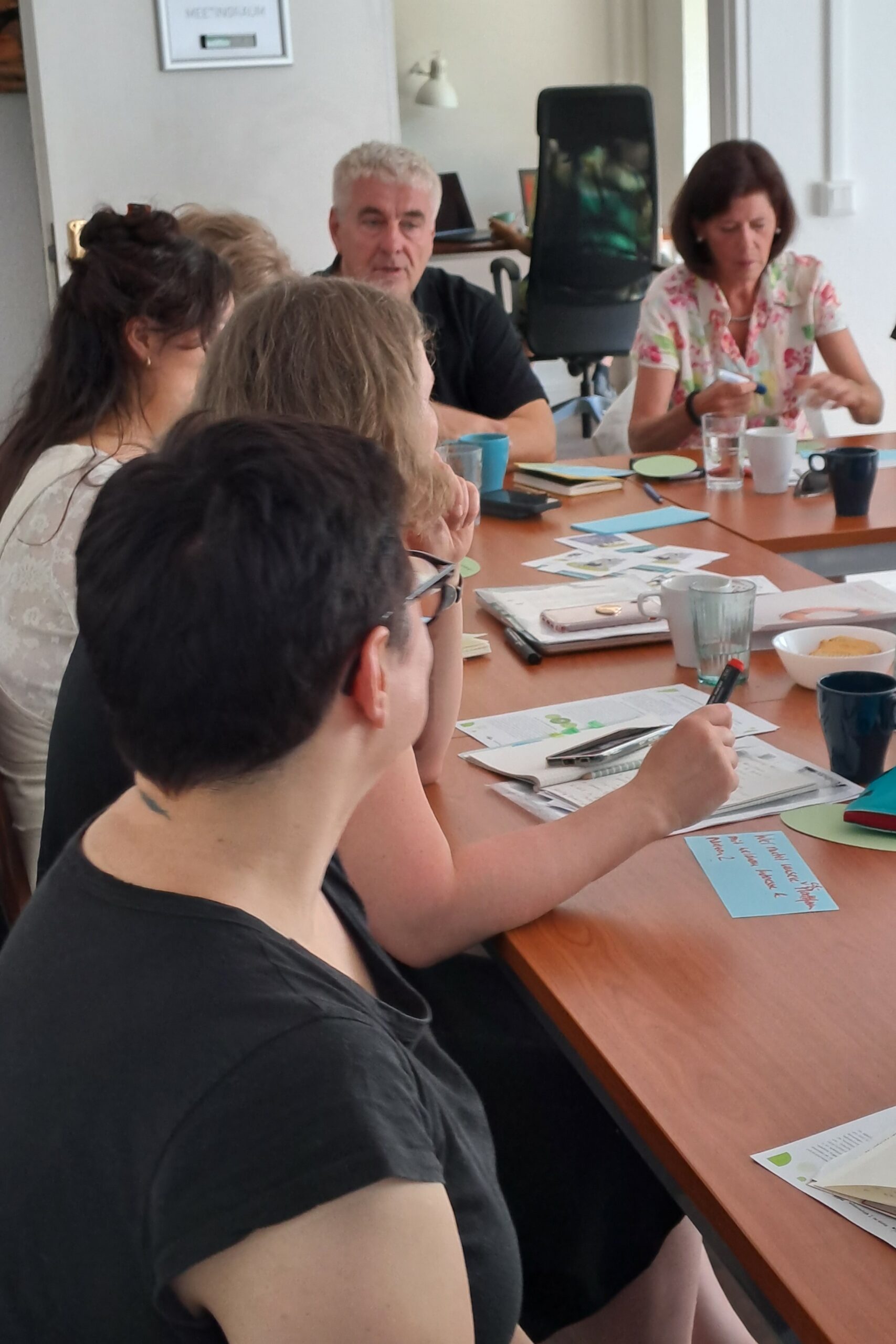 Teilaufnahme eines Workshops, in dem sechs Personen am Tisch sitzen und eine angeregte Diskussion führen. Auf dem Tisch liegen Notizen und Moderationskarten.