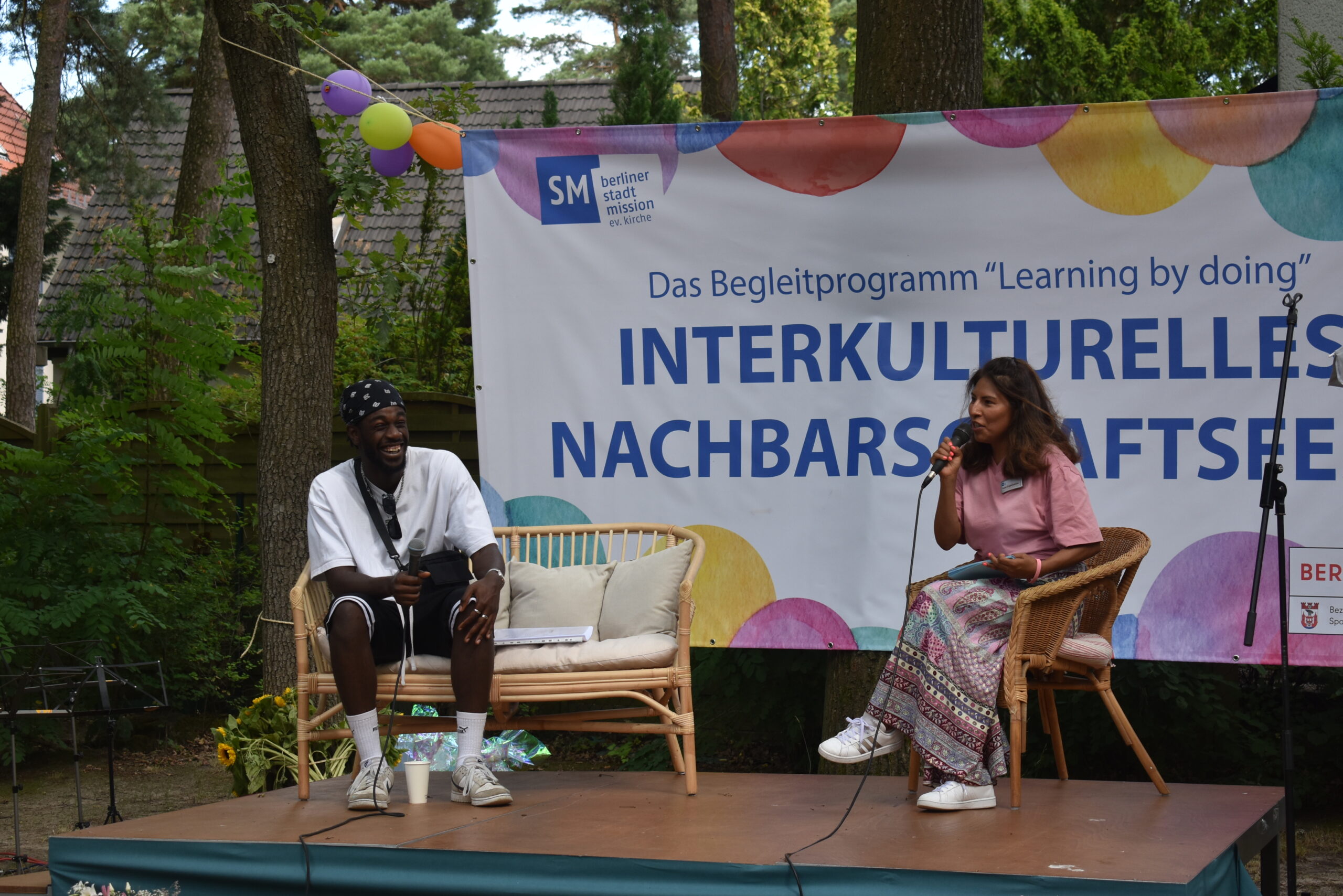 Auf einer Bühne sitzen zwei Personen, ein Mann und eine Frau, die auf einem Fest sprechen. Im Hintergrund ist ein großes Banner mit dem Schfriftzug: Das Begleitprogramm "Learning by doing", Interkulturelles Nachbarschaftsfest