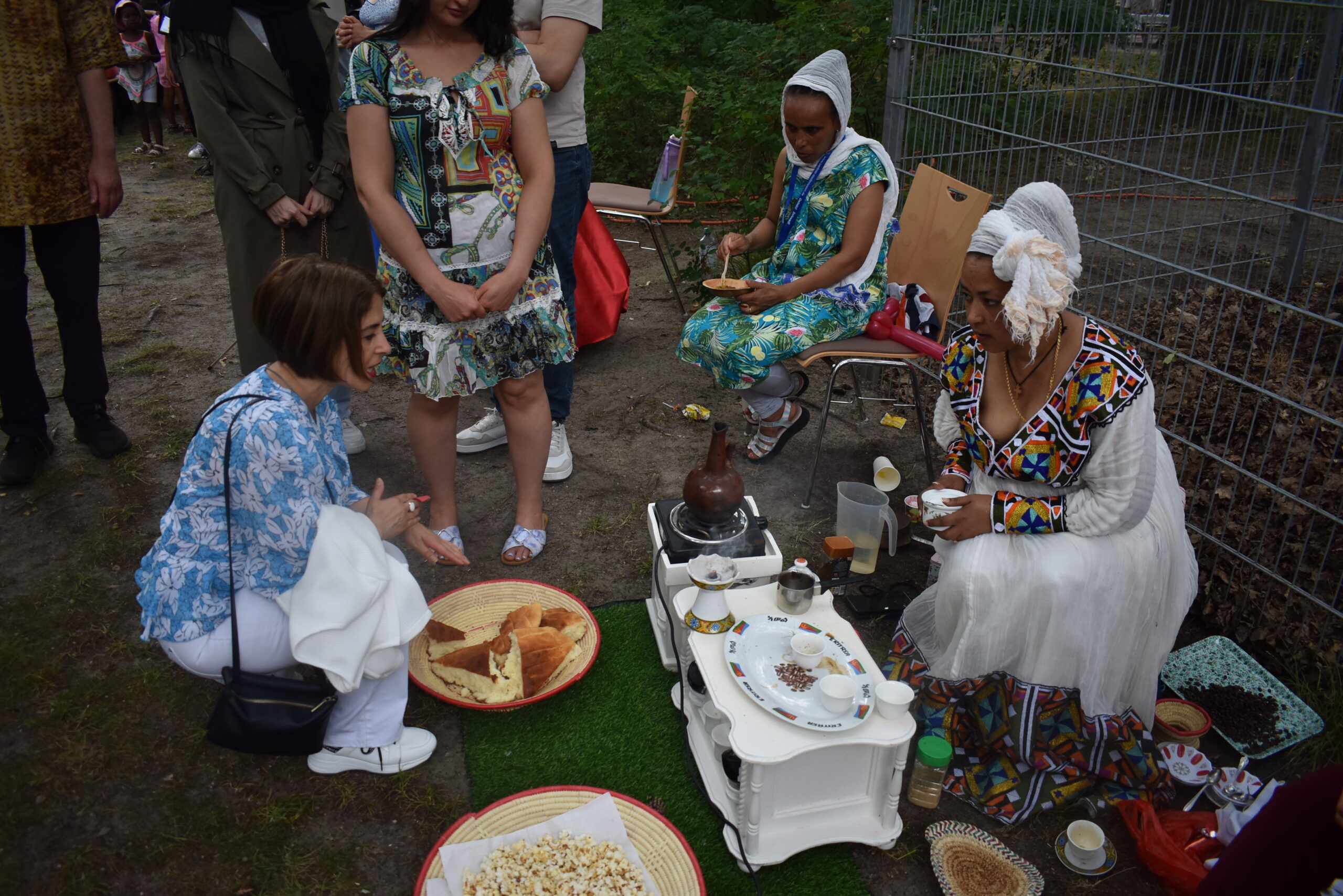 Auf einem Fest sitzen zwei Frauen und bieten traditionelles afrikanisches Essen an. Eine weitere Frau spricht mit ihnen.