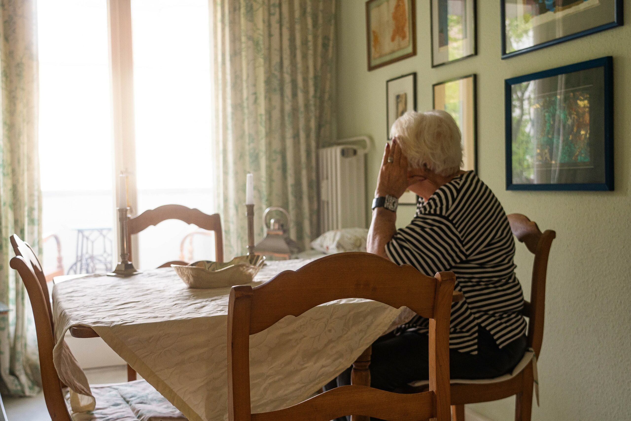 Eine ältere Frau sitzt allein an einem mit weißer Tischdecke bedeckten Esstisch. Sie stützt den Kopf mit einer Hand und blickt aus dem Fenster. An den Wänden hängen mehrere gerahmte Bilder.