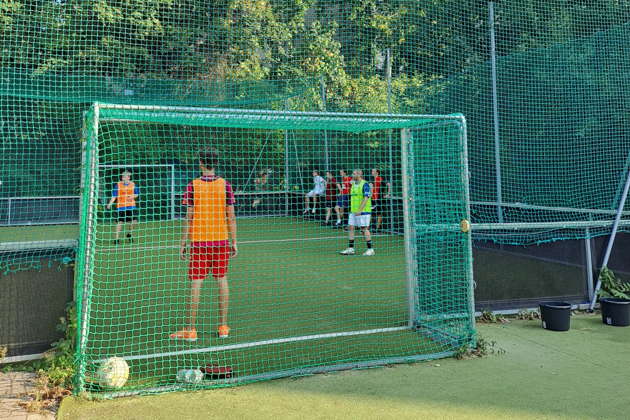 Ein kleines Fußballfeld mit Kunstrasen, aus der Perspektive hinter dem Tor aufgenommen. Auf dem Spielfeld sind sieben Spieler verteilt zu sehen.