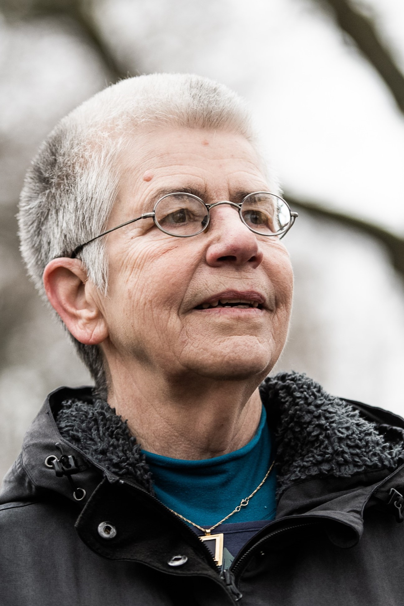 Eine ältere Dame mit kurzen grau-weißen Haaren, Brille, einem schwarzen Anorak und einer goldenen Kette schaut in die Ferne.