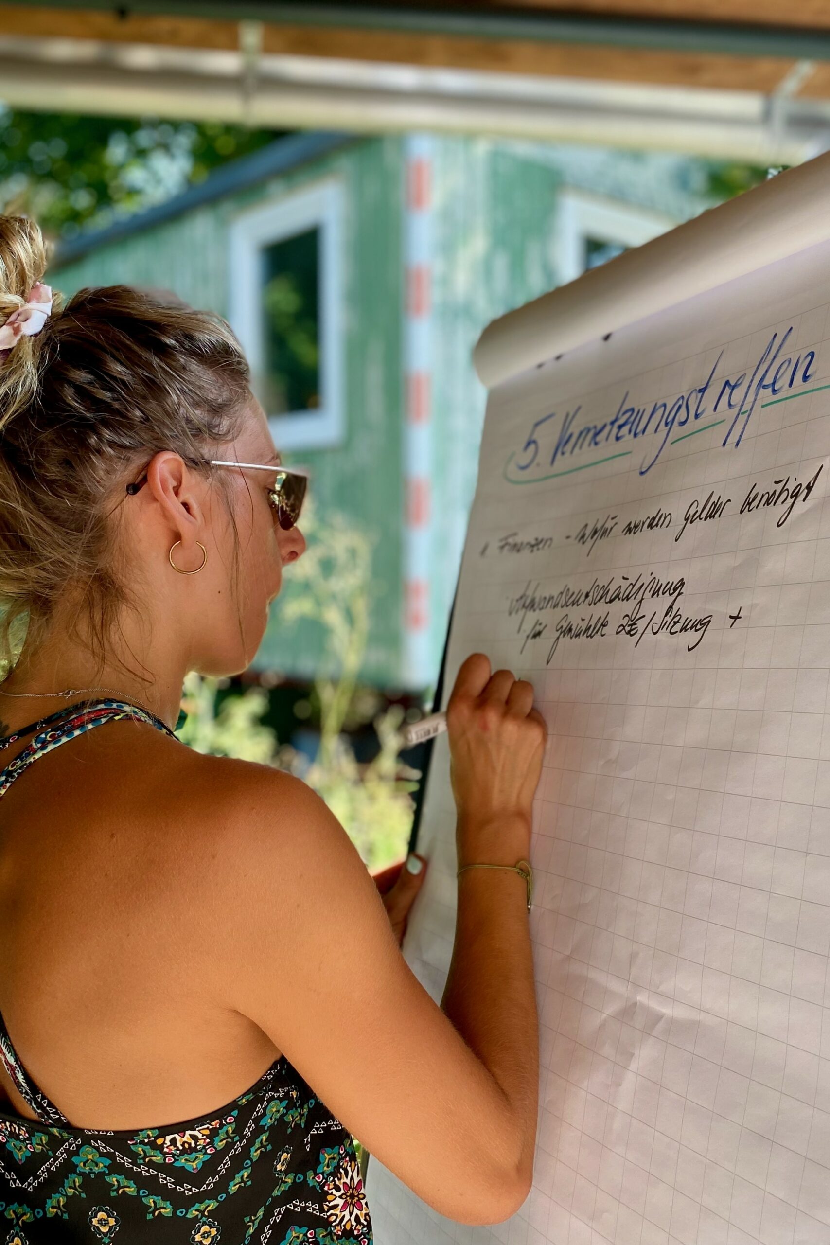 Eine Frau mit blonden Haaren steht an einer Flipchart und schreibt mit einem schwarzen Edding Punkte eines Treffens auf.