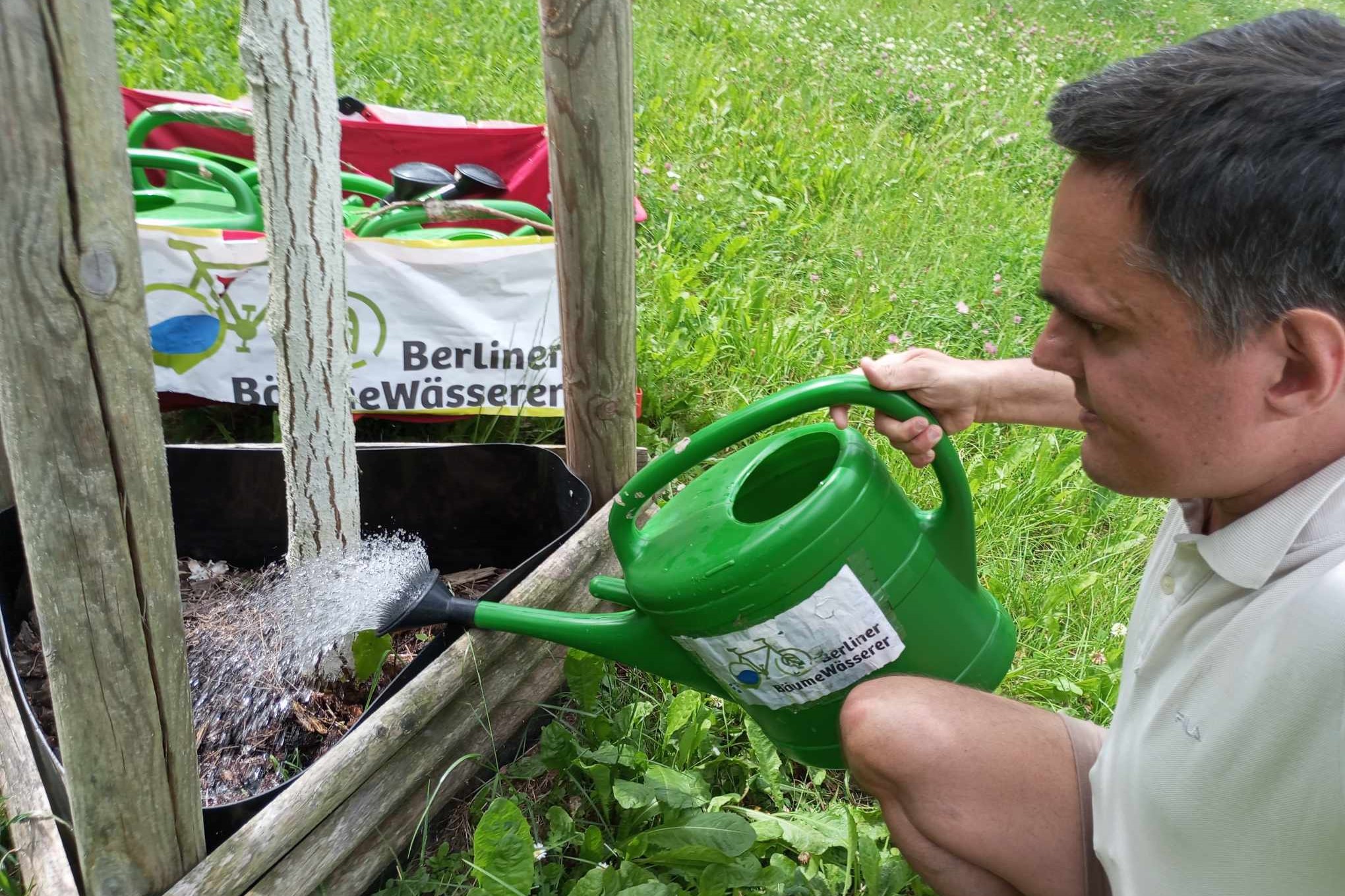 Ein Mann mit kurzen braunen Haaren und einem weißen Polo-Shirt gießt mit einer grünen Gießkanne einen Baum. Im Hintergrund sind weitere Gießkannen zu sehen.