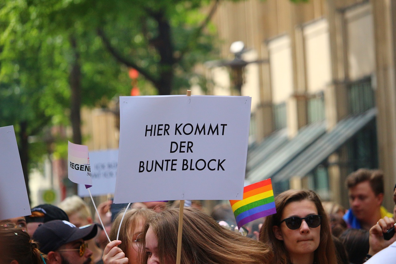 Im Vordergrund steht eine Demonstrantin, die ein Plakat mit dem Schriftzug "Hier kommt der bunte Block" hochhält, während im Hintergrund die Demonstration weitergeht.