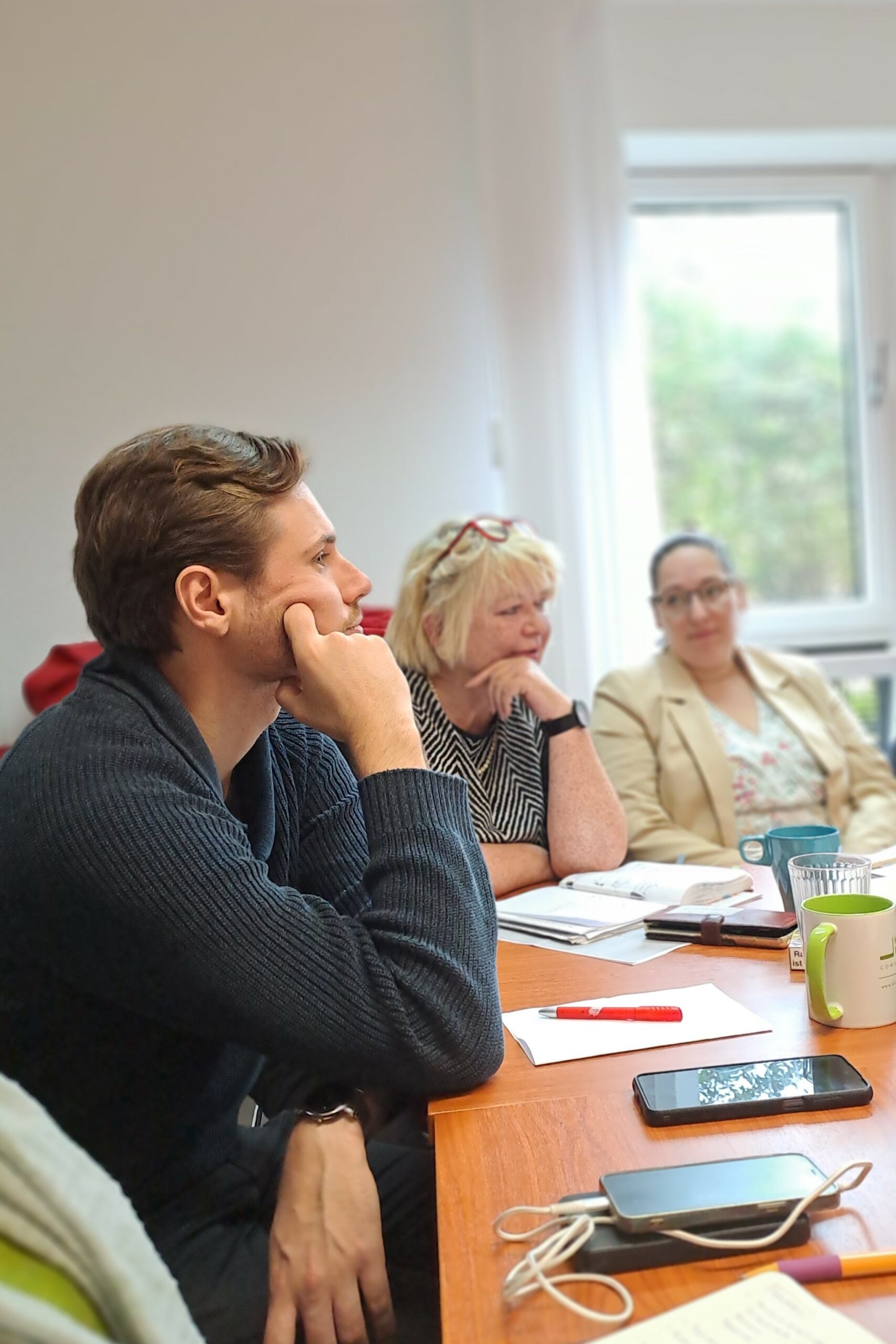 Ein junger Mann sitzt an einem Tisch, stützt seinen Kopf mit einer Hand und scheint zuzuhören. Im Hintergrund sind zwei Frauen an einem weiteren Tisch zu sehen.