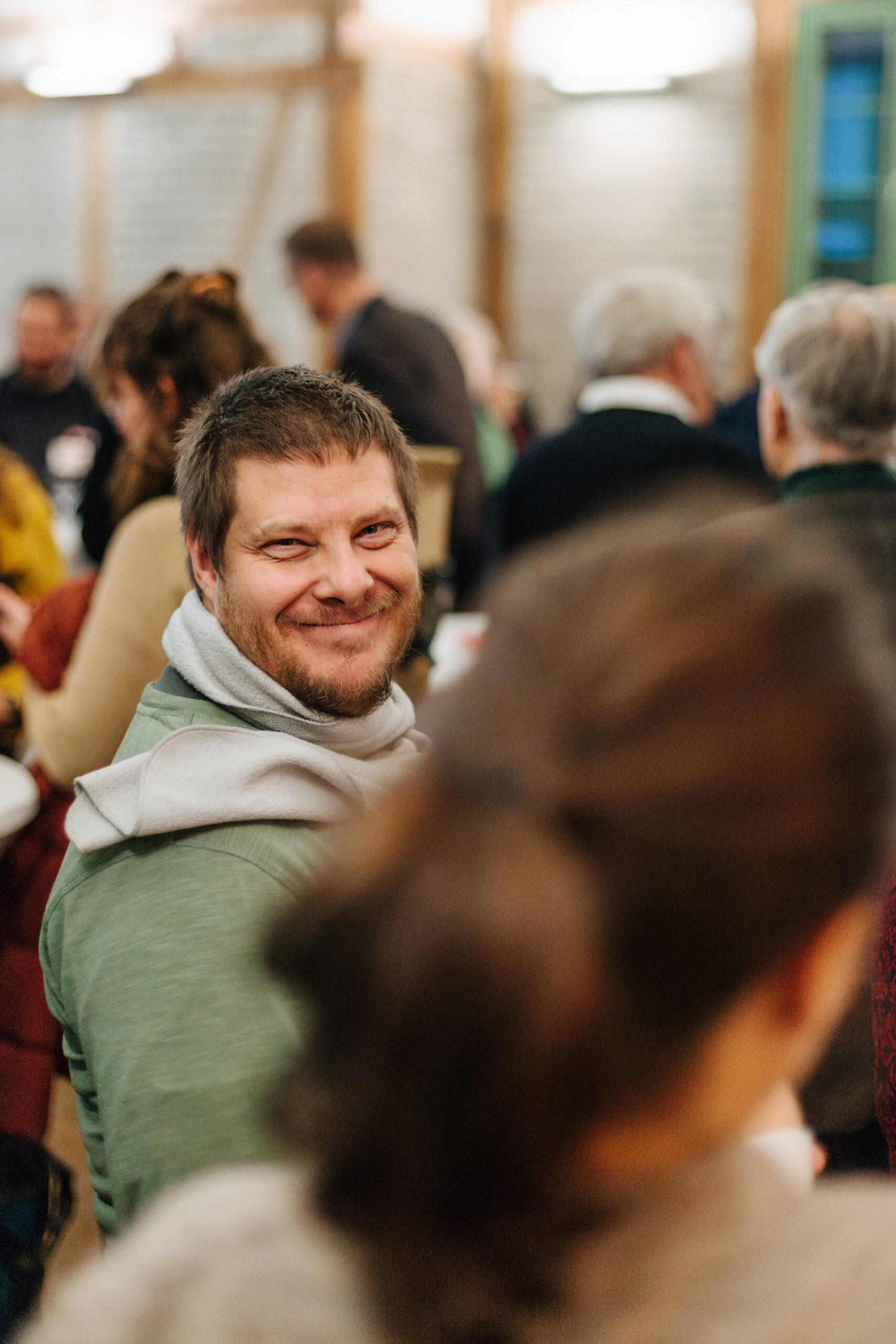 Ein Mann mit grünem Pullover und hellem Schal lächelt während einer Veranstaltung in die Kamera.