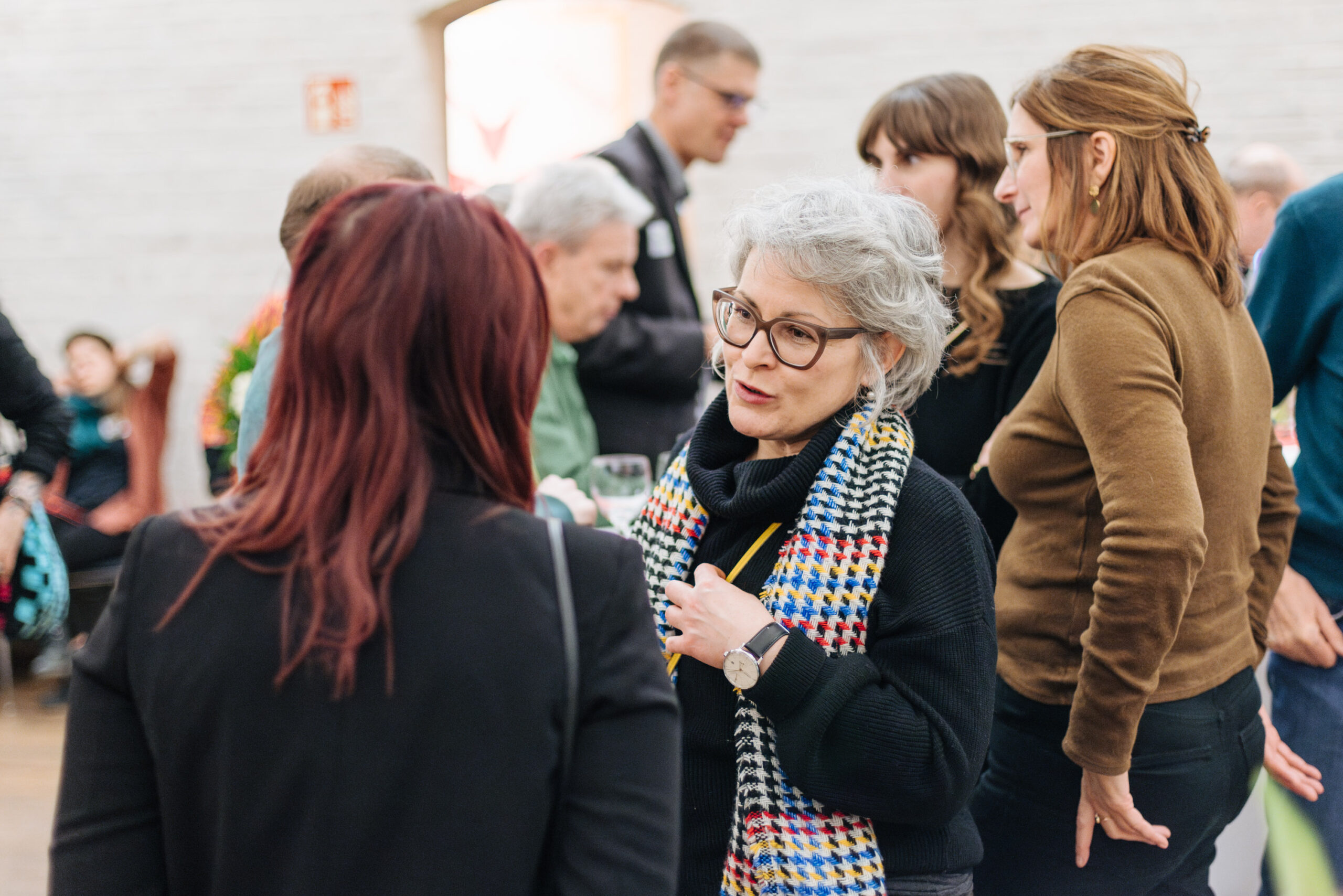 Zwei Frauen unterhalten sich angeregt am Rand einer Veranstaltung. Im Hintergrund sind weitere Gäste zu sehen.